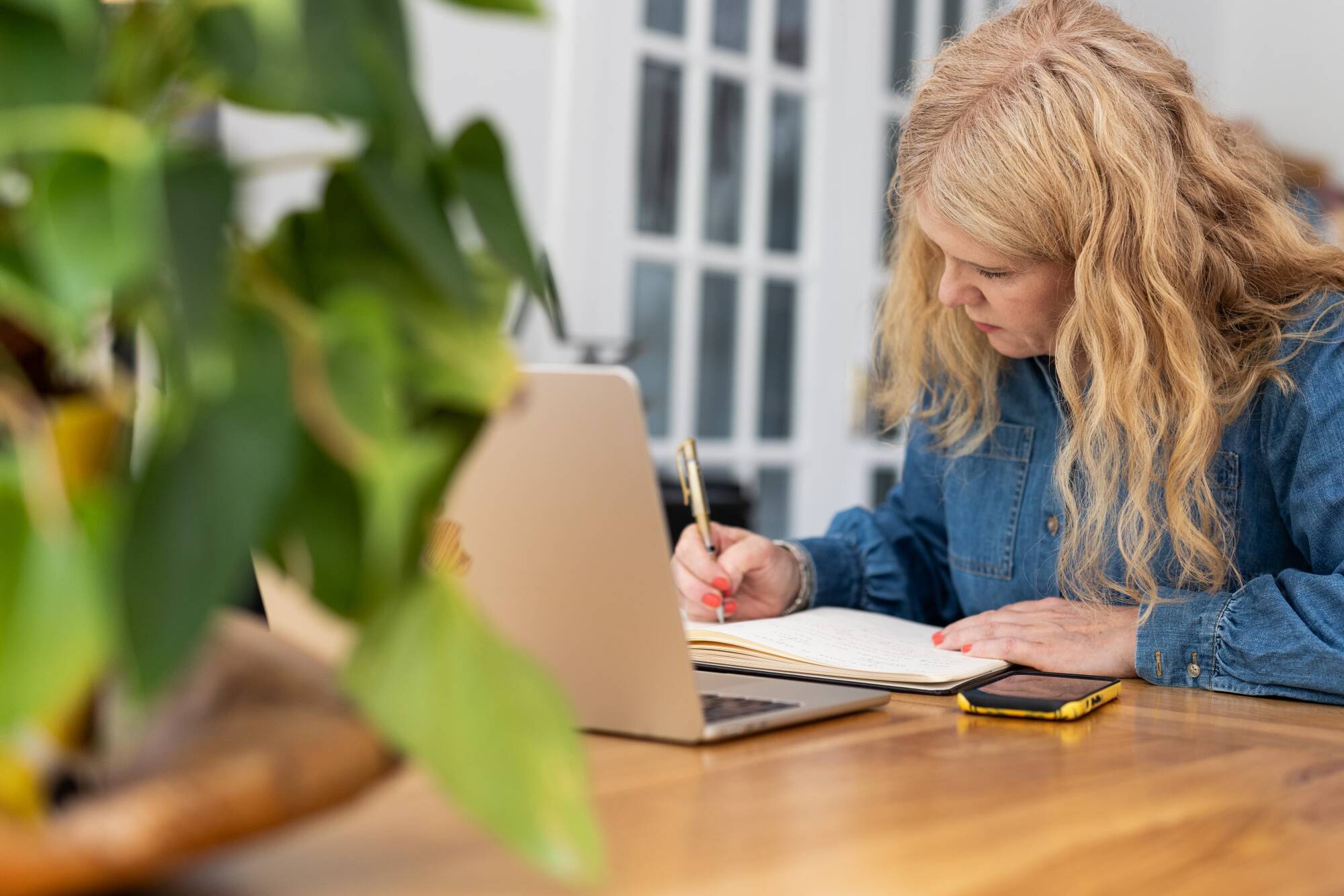 A woman with long blond hair sits at a wooden table, writing in a notebook beside an open laptop and smartphone in a bright home office, with a blurred green plant in the foreground.